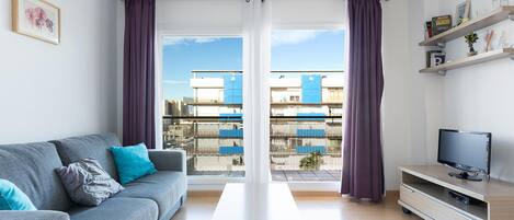 A modern living room with a grey sofa, a wooden coffee table, a TV on a wooden stand, and a balcony with purple curtains.