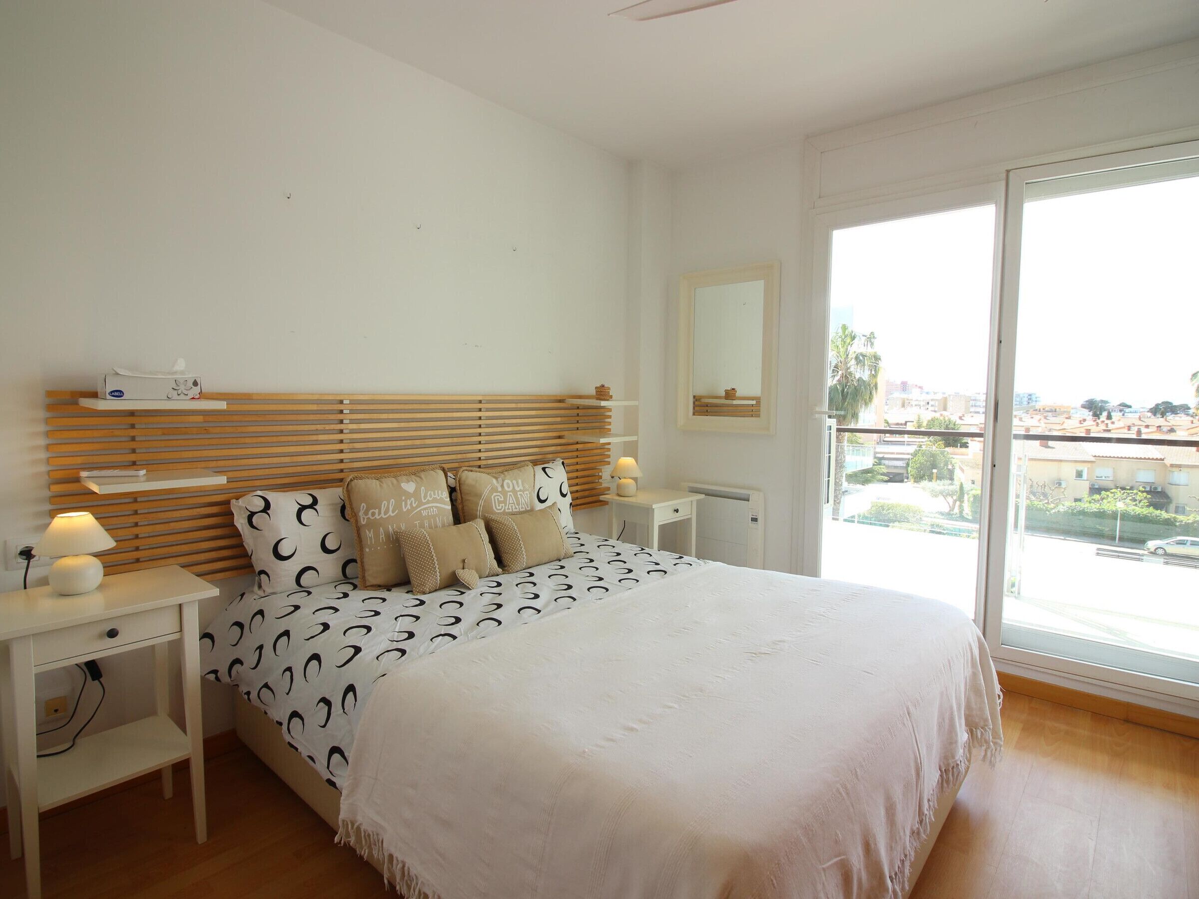 Un dormitorio con cama, mesitas de noche, cómoda, espejo y un gran ventanal con vistas a edificios y palmeras.
