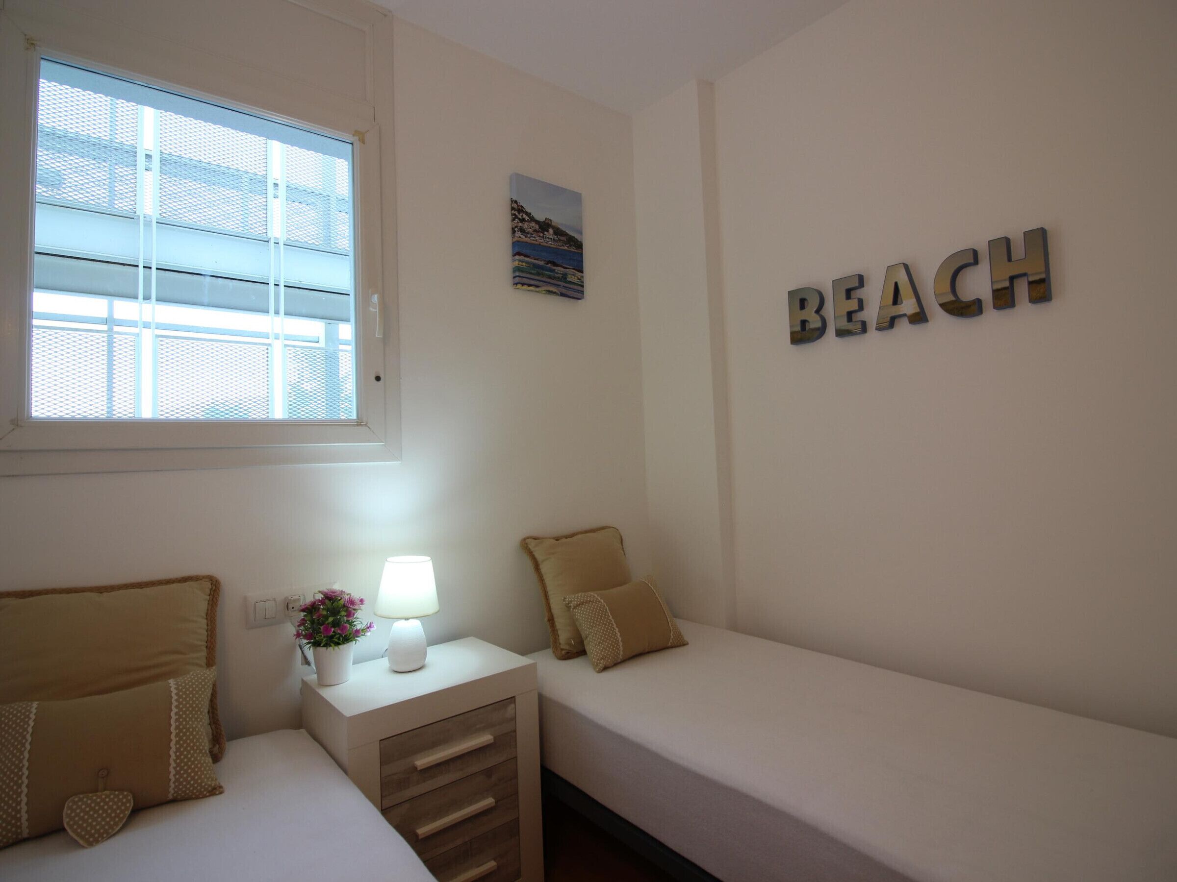 Une chambre avec une fenêtre, une table de chevet avec une lampe, deux lits et le mot «PLAGE» écrit sur le mur.