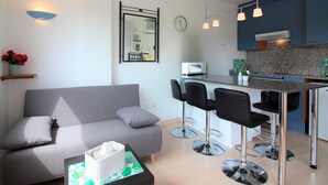 Ein modernes Wohnzimmer mit einem grauen Sofa, einem kleinen Tisch mit Taschentuchbox und einer Küchenzeile mit Mikrowelle und Barhocker.