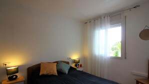 Ein Schlafzimmer mit einem Bett, zwei Nachttischen, einer Lampe auf jedem Tisch, einem Fenster mit Vorhängen und einem runden, an der Wand befestigten Spiegel.