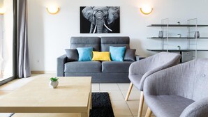 Ein modernes Wohnzimmer mit einem grauen Sofa, einem Couchtisch und einem schwarzen Teppich. An der Wand hängt ein Elefantenbild, und eine Metallregalvorrichtung zeigt Pflanzen.