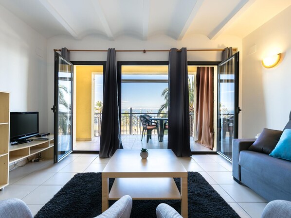 A modern living room with a flat-screen TV, a sofa, a coffee table, and a balcony with a view of the sea.