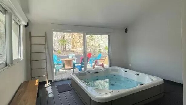 A small room with a hot tub, a wooden bench, and a sliding glass door leading to an outdoor area with chairs and a table.