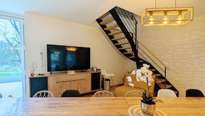 A modern living room with a wooden dining table, a large flat-screen TV, a staircase with a black railing, and a white brick wall.