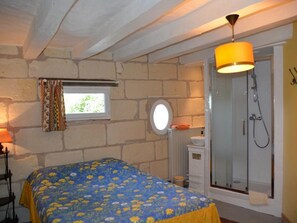 Un dormitorio con cama, ventana con cortinas, espejo redondo, ducha y lámpara.