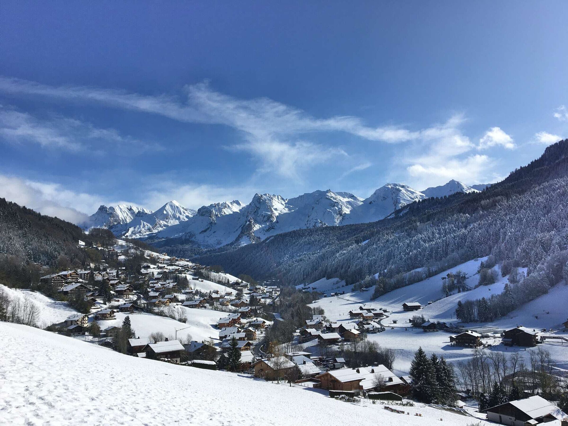 Ein verschneites Dorf mit traditionellen Holzhäusern, einem Berg im Hintergrund und blauem Himmel.