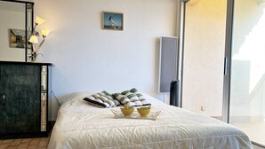 Quarto com cama, almofadas, mesa de cabeceira com abajur, pintura e vista através de uma porta de vidro.