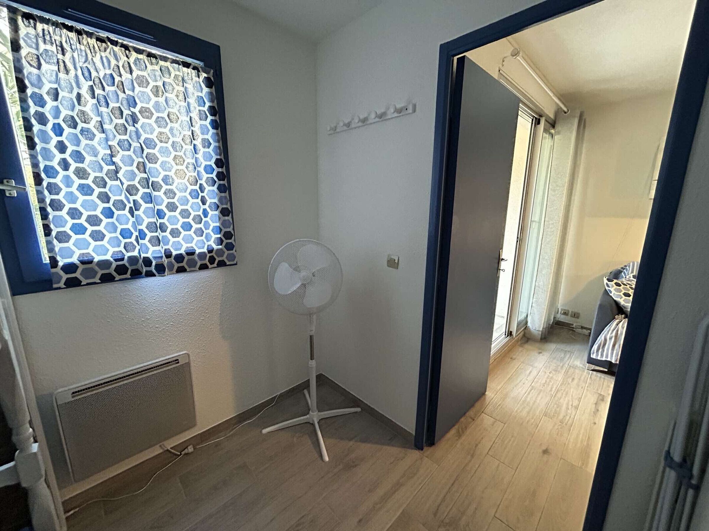 Ein Zimmer mit einem Fenster, das mit gemusterten Vorhängen verziert ist, einem Standventilator und einer Tür zu einem weiteren Raum.