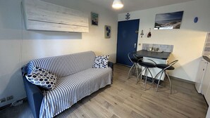 Een compacte woonruimte met een gestreepte bank, een eettafel met twee stoelen, een blauwe deur en een klein keukentje.