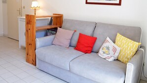 A grey sofa with four pillows, a wooden side table, a lamp, and a framed painting on the wall. - Sunny 2-Room Apartment with Terrace and Pool, Near Beach (Le Lavandou)