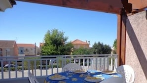 Un balcón con una mesa dispuesta para cuatro personas, sillas de plástico blancas y un mantel azul con estampados amarillos.