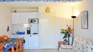 Cocina compacta con armarios blancos, microondas, cafetera y lavadora. Hay una mesa de comedor con sillas y un sofá estampado con motivos florales.