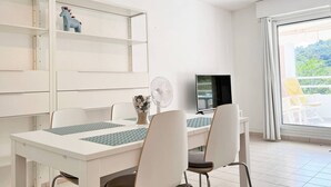 Ein moderner Essbereich mit einem weißen Tisch, Stühlen und einem Ventilator. Auf dem Tisch steht ein Fernseher und ein Fenster bietet Blick auf Grünflächen.