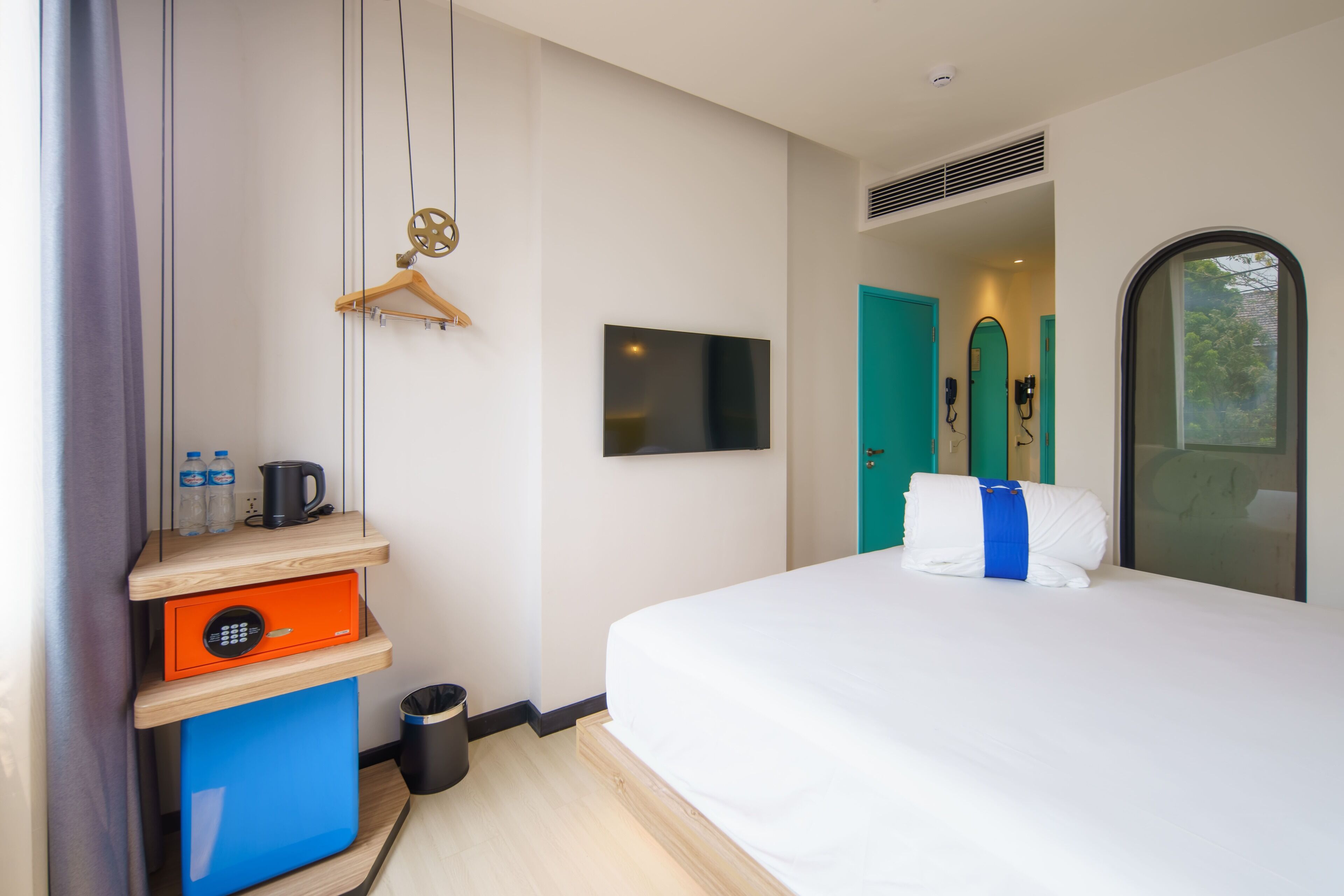 A hotel room with a bed, a TV, a wardrobe, a small desk with a kettle, and a trash bin.