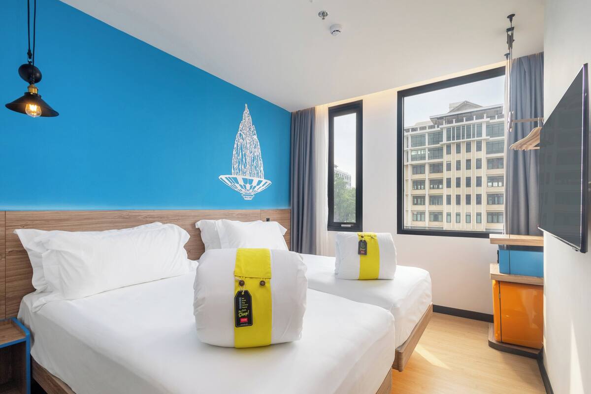 Een hotelkamer met twee bedden, een blauwe accentmuur, een groot raam en uitzicht op een gebouw.