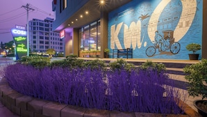 A hotel exterior with a large mural, a neon sign, and a landscaped area with purple plants. - COSI Vientiane Nam Phu (Vientiane)