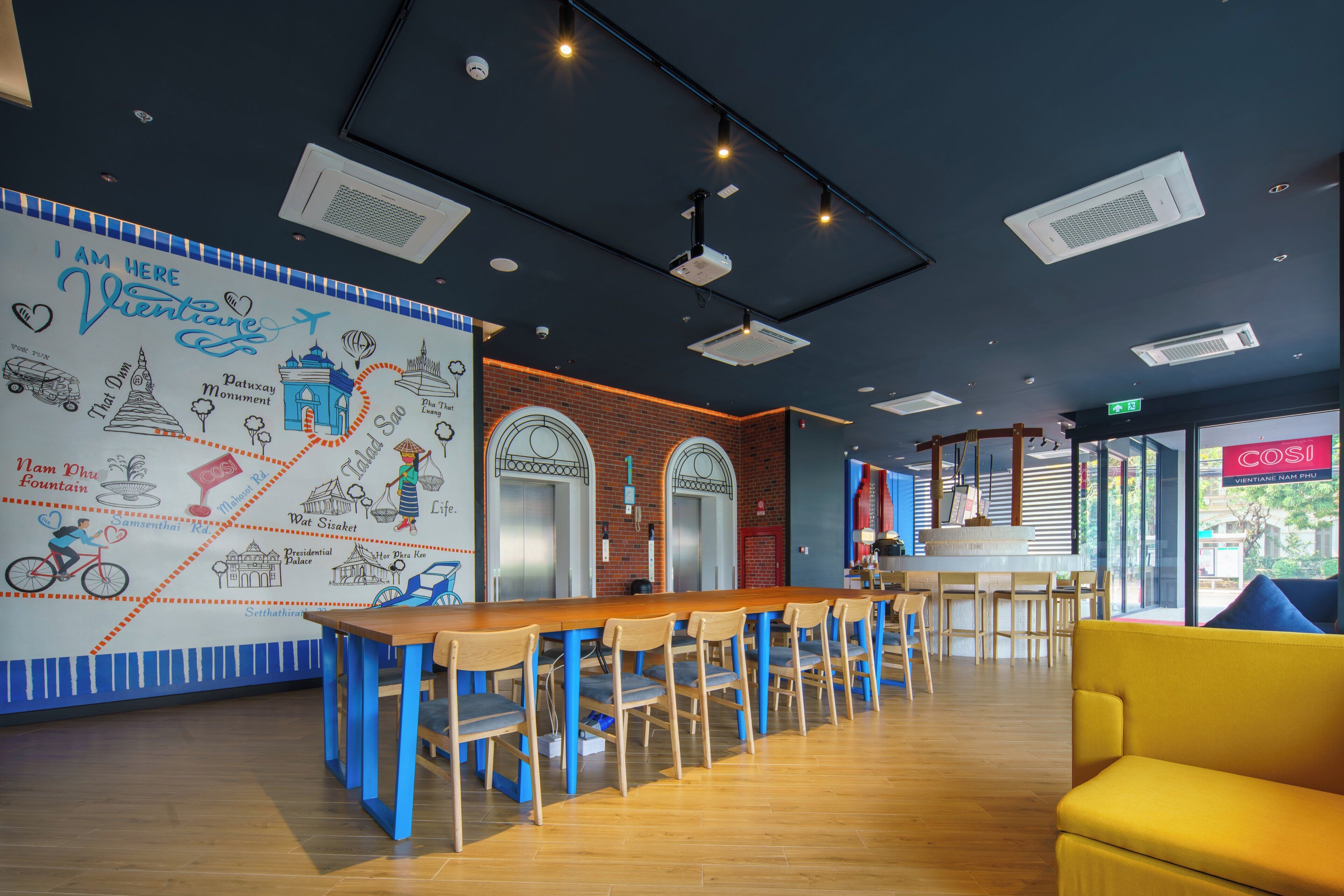 A modern bar area with a wooden counter and blue chairs, a yellow sofa, and a wall with travel-themed illustrations.