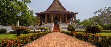 Un temple thaïlandais traditionnel, avec un chemin dallé menant à l’entrée, entouré de jardins bien entretenus et sous un ciel dégagé.