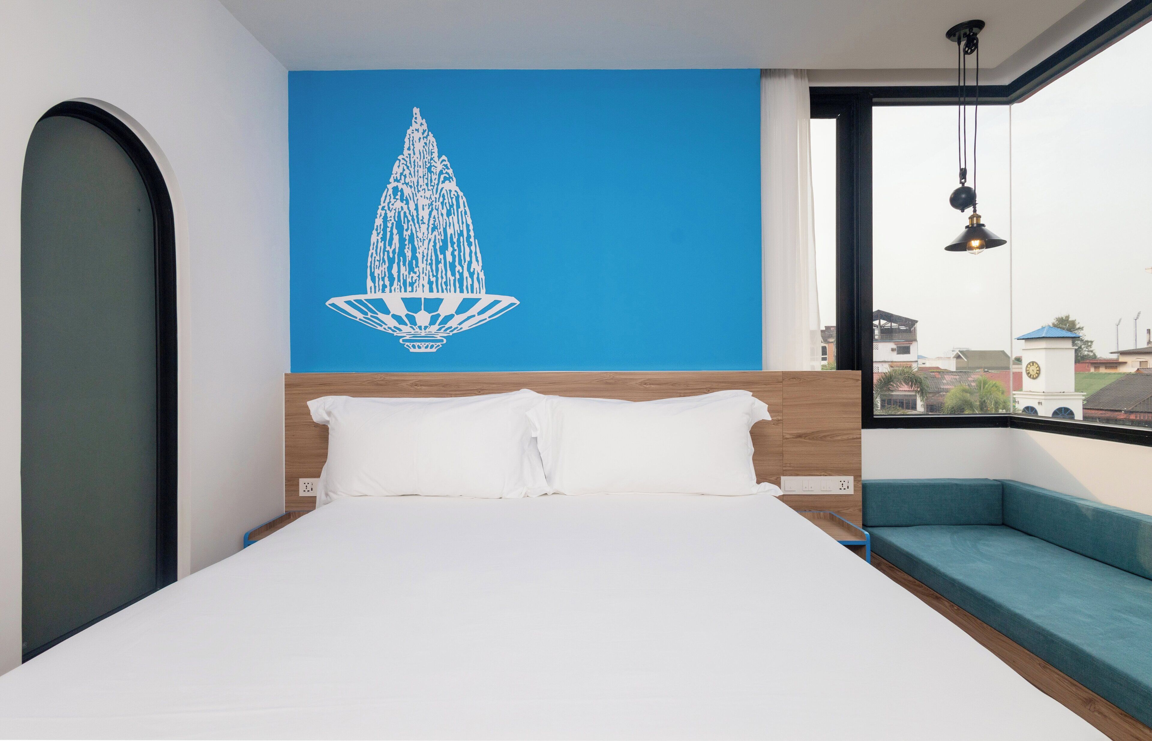 Un hotel moderno con una cama grande, una pared azul con una obra de arte de una fuente blanca, vista de la ciudad a través de un ventanal grande y un sofá azul junto a la ventana.