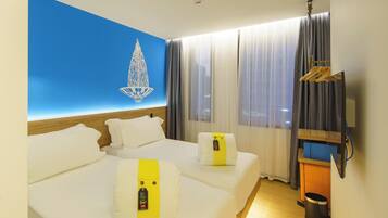 Habitación de hotel con dos camas, pared azul con candelabro, cabecera de madera, televisor y ventana con cortinas.