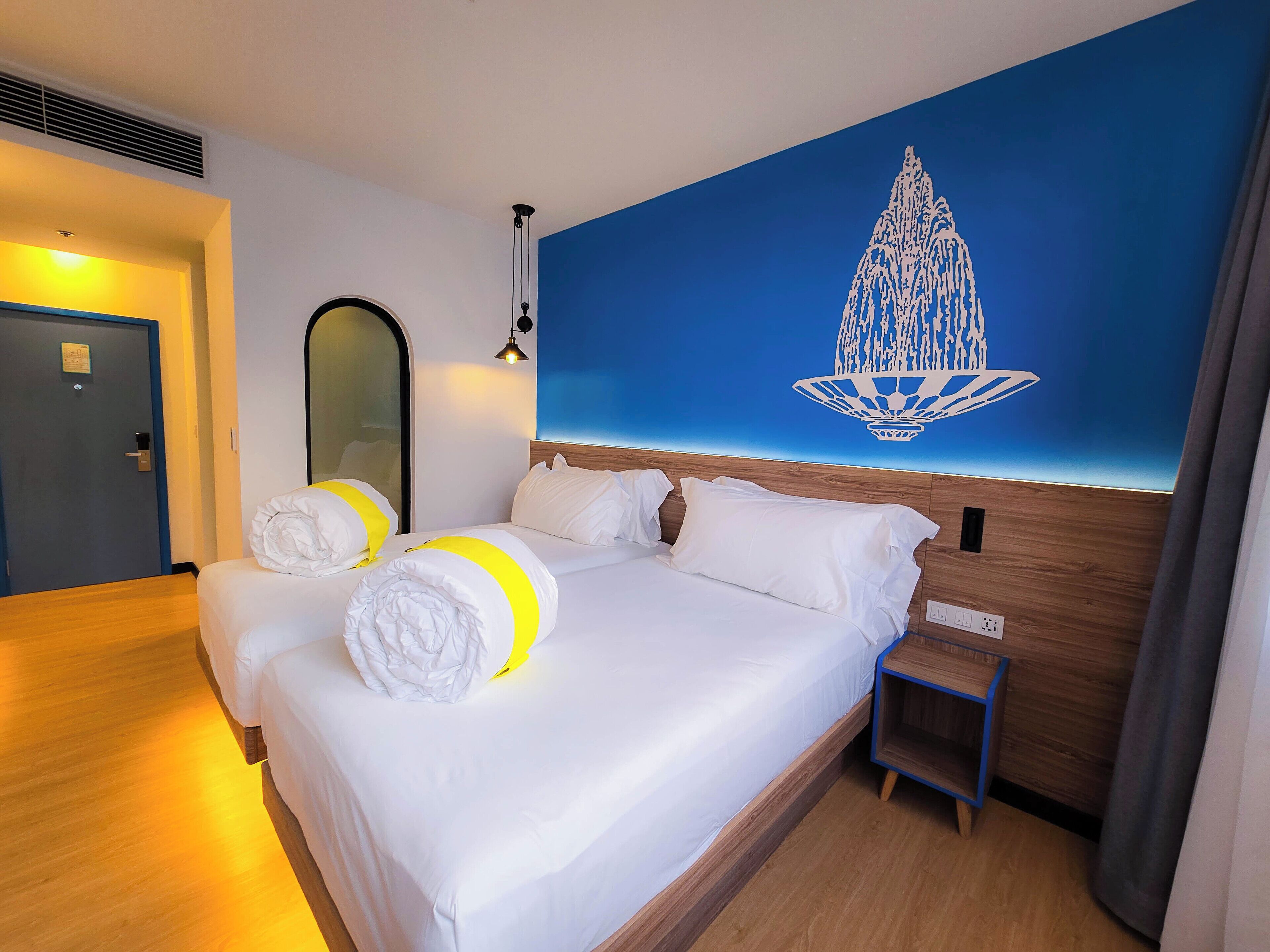A hotel room with a bed, a nightstand, a mirror, and a wall with a decorative light fixture.