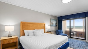 A bedroom with a large bed, a nightstand, a lamp, a chair, and a view of the beach. - Oceanfront Studio with Private Porch 1st Floor (Pawleys Island)