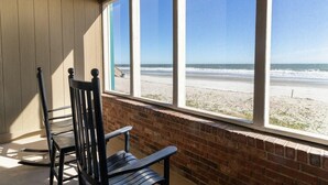 A room with a view of the beach through large windows, a brick wall, and a wooden chair. - Oceanfront Studio with King Bed and Porch 1st Fl (Pawleys Island)