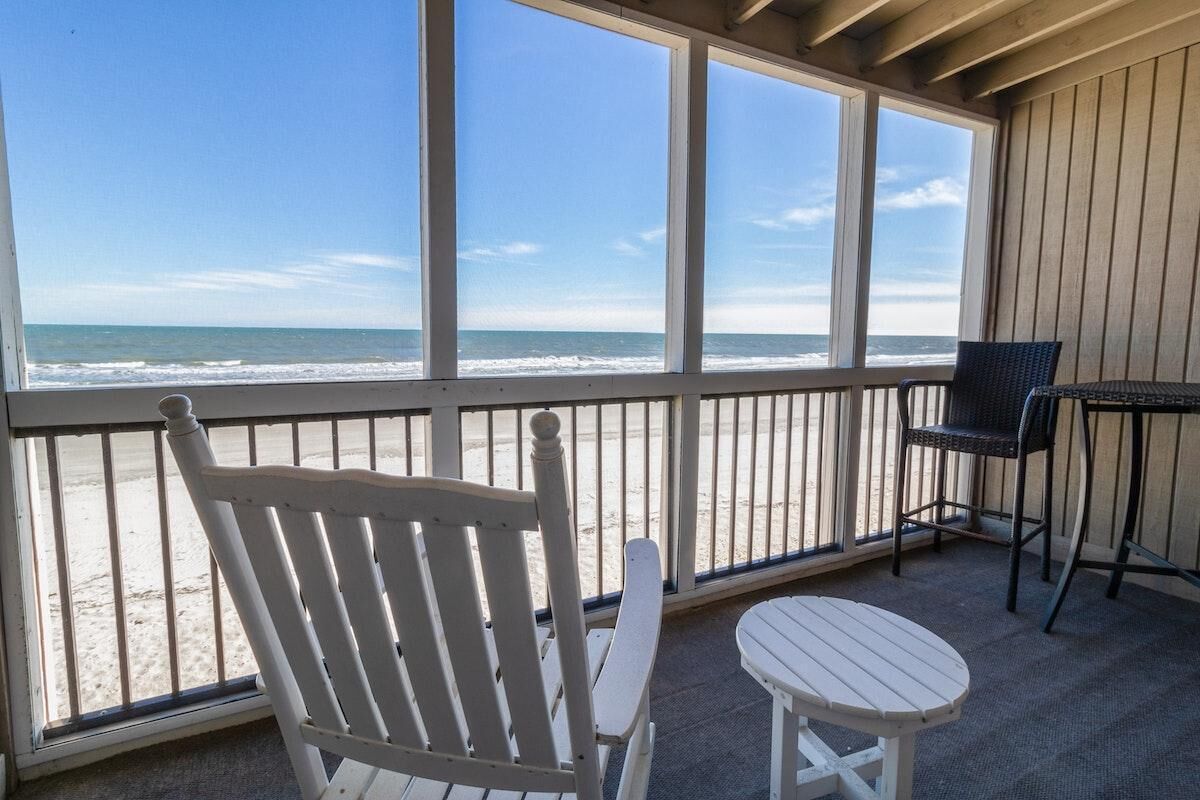 Ein Balkon mit weißem Stuhl und Tisch, der Blick auf einen Strand mit Wellen bietet.