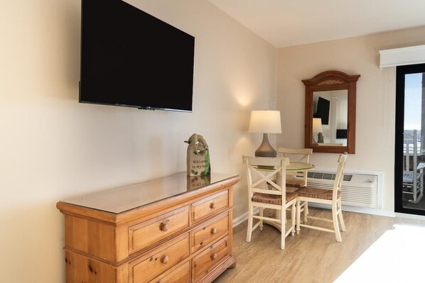 A room with a wooden dresser, a flat-screen TV mounted on the wall, a table with chairs, a lamp, and a mirror.