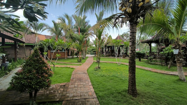 Un chemin de jardin qui mène à un bâtiment doté d’un toit en tuiles, entouré de palmiers et d’une végétation luxuriante.