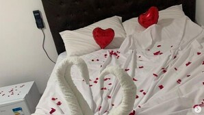 A bed with a white duvet and red heart-shaped pillows, a bedside table with a tissue box, and a power outlet on the wall. - Pousada Brisa Suítes (Ipojuca)