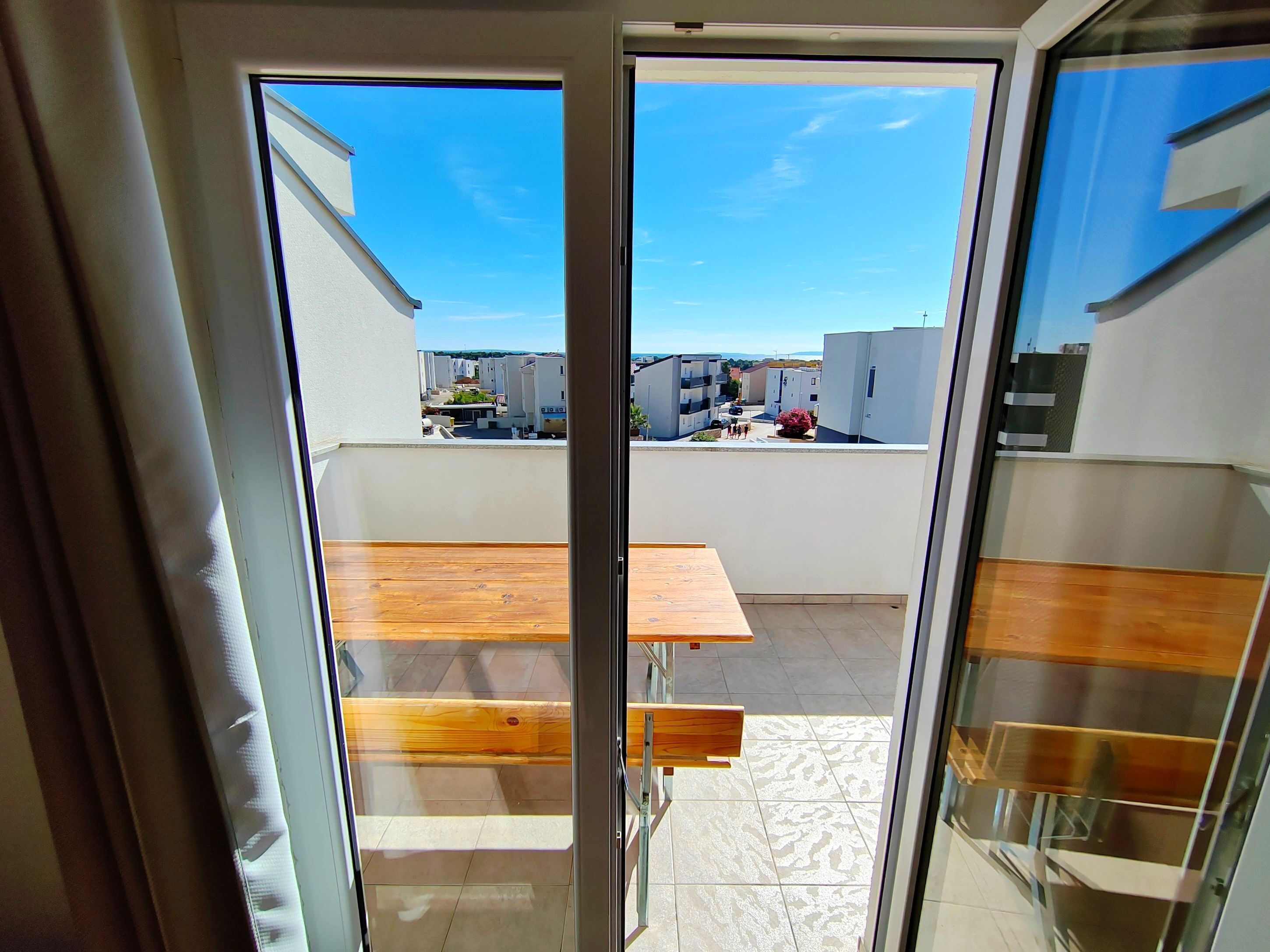 Uitzicht door een glazen deur op een terras met een houten tafel en banken, een betegelde vloer en een helderblauwe lucht.