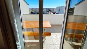 A view through a glass door to an outdoor patio with a wooden table and benches, a tiled floor, and a clear blue sky.