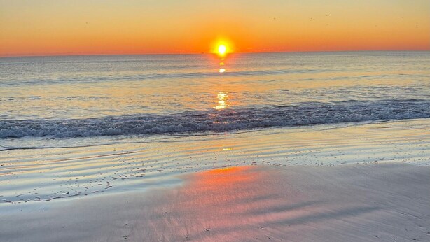 Ein Strand bei Sonnenuntergang, die Sonne tief am Horizont, ihr Licht spiegelt sich im Wasser.