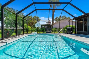 A swimming pool with a basketball hoop, surrounded by a covered area with a view of trees and a building.