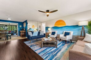 Un salon moderne avec un ventilateur de plafond, un grand tableau représentant un coucher de soleil et une vue à travers une porte vitrée.