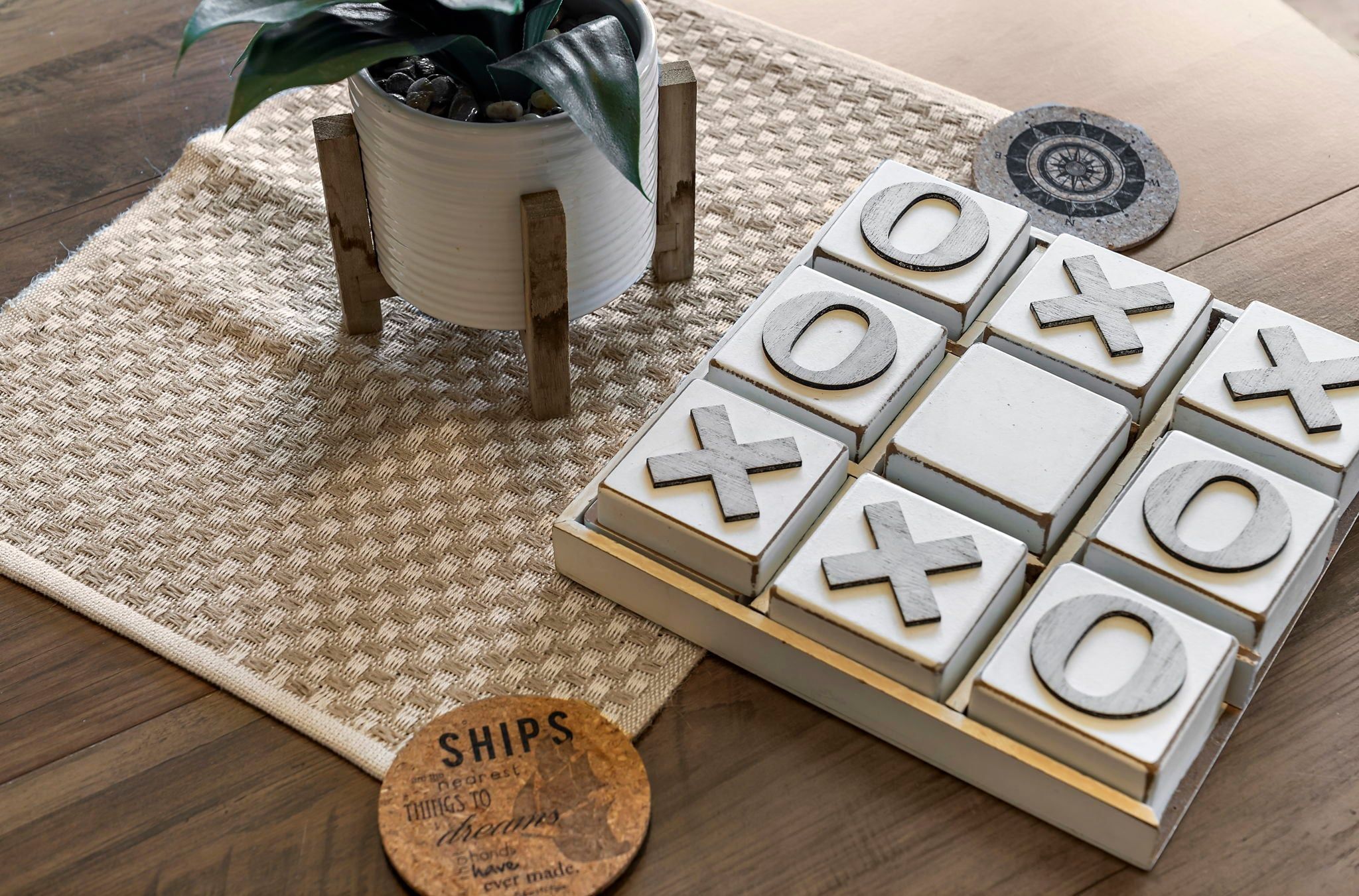 Una mesa de madera con un tapete tejido, una planta en maceta, una cruz de madera, una brújula y un tablero de juego de madera con símbolos de X y O.