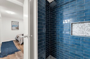 Une salle de bain avec des carreaux bleu foncé, un espace douche et une porte donnant sur une chambre à coucher équipée d’un lit et d’un tapis.
