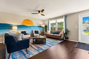A living room with a ceiling fan, a large painting of a sunset, and a view of a house through the window. - ⚓Sunset Getaways! Private Heated Pool home, Cinema/Arcade Room⚓ (Bradenton)