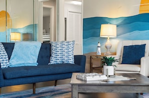 A living room with a blue sofa, a white armchair, and a coffee table. There is a lamp on a side table, a potted plant, and a wall with a painted ocean scene. - ⚓Sunset Getaways! Private Heated Pool home, Cinema/Arcade Room⚓ (Bradenton)