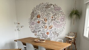 Een eethoek met een houten tafel, witte stoelen, een geweven mand en een muurschildering.
