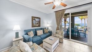 Een woonkamer met een blauwe bank, twee fauteuils, een salontafel, een plafondventilator en een groot raam met uitzicht op het strand.