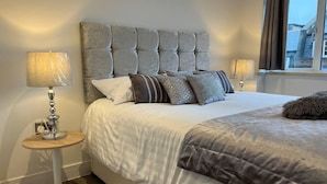 Una cama bien hecha con un tablero de cabecera tapizado en gris, dos lámparas de noche y un almohadón mullido.