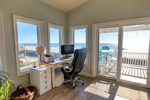 A room with a desk, chair, and large windows overlooking the beach.