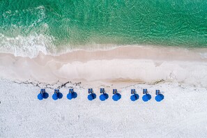 An aerial view of a beach with white sand and turquoise water. There are ten blue umbrellas lined up in a row on the sand.