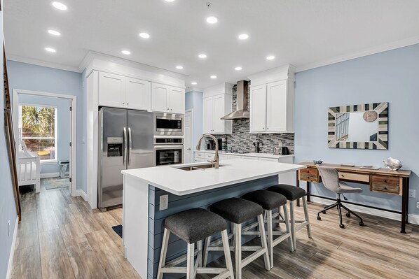 A modern kitchen with white cabinets, stainless steel appliances, a large island with bar stools, and a wooden desk with a chair.