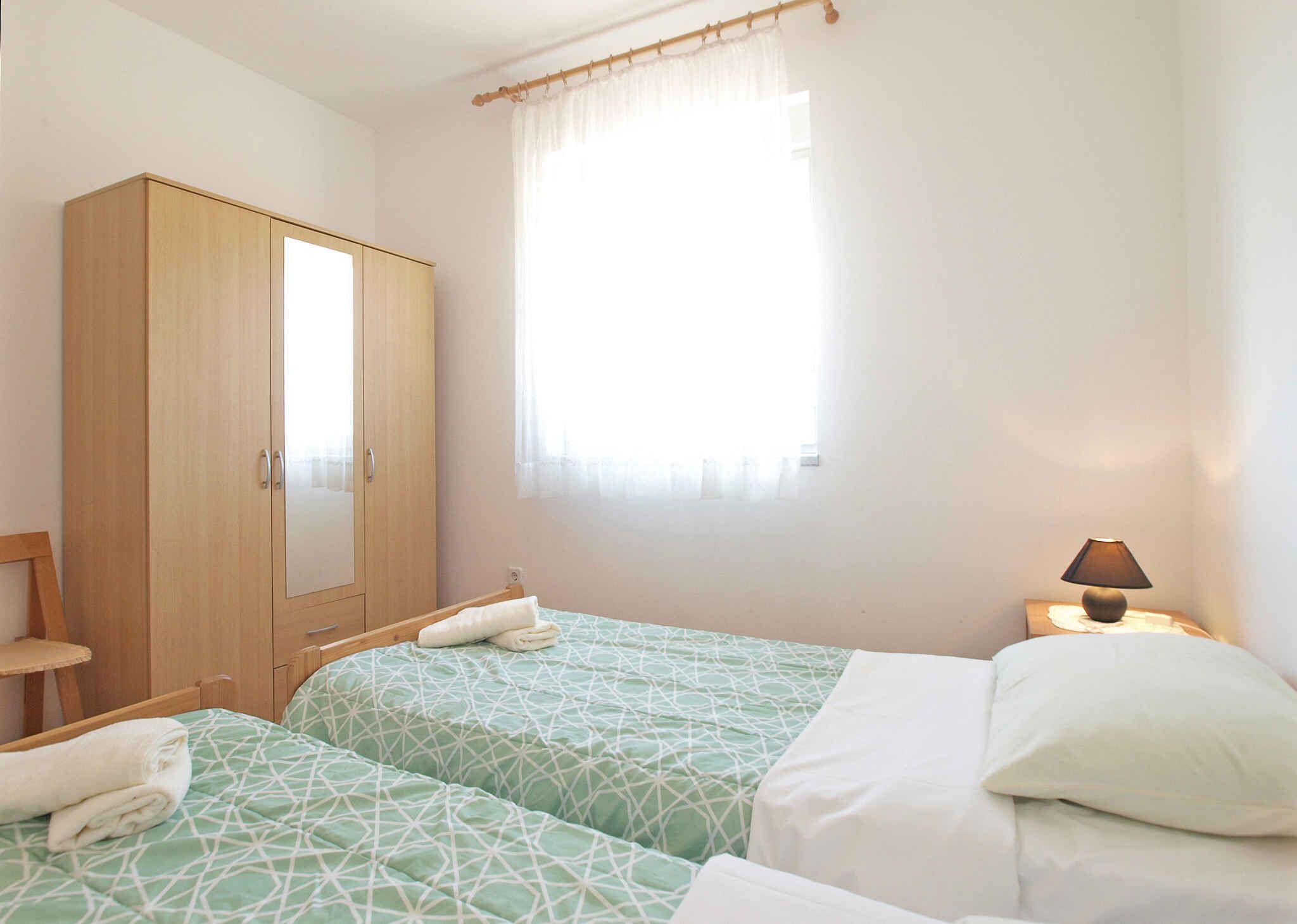 A bedroom with two beds, a wardrobe, a bedside table with a lamp, and a window with sheer curtains.