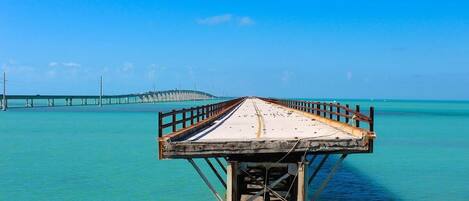 A pier extending into a clear blue sea with a distant bridge.