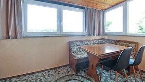 Ein Zimmer mit Holzboden, einem großen Fenster mit Vorhängen, einem Holztisch, zwei Stühlen und einer Sitzbank mit gemustertem Bezug.